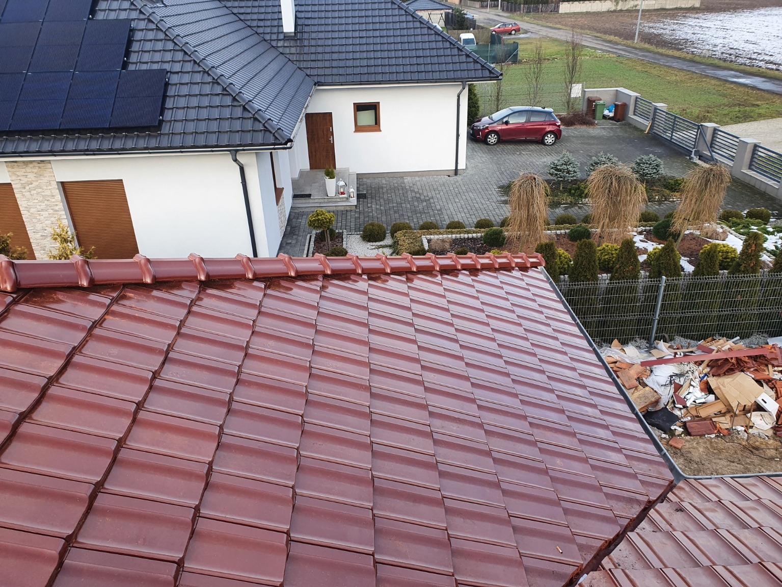 Nowe, błyszczące dachówki w kolorze ceglanej czerwieni, ułożone na dachu nowoczesnego domu z panelami słonecznymi, widoczne pozostałości po starej dachówce.