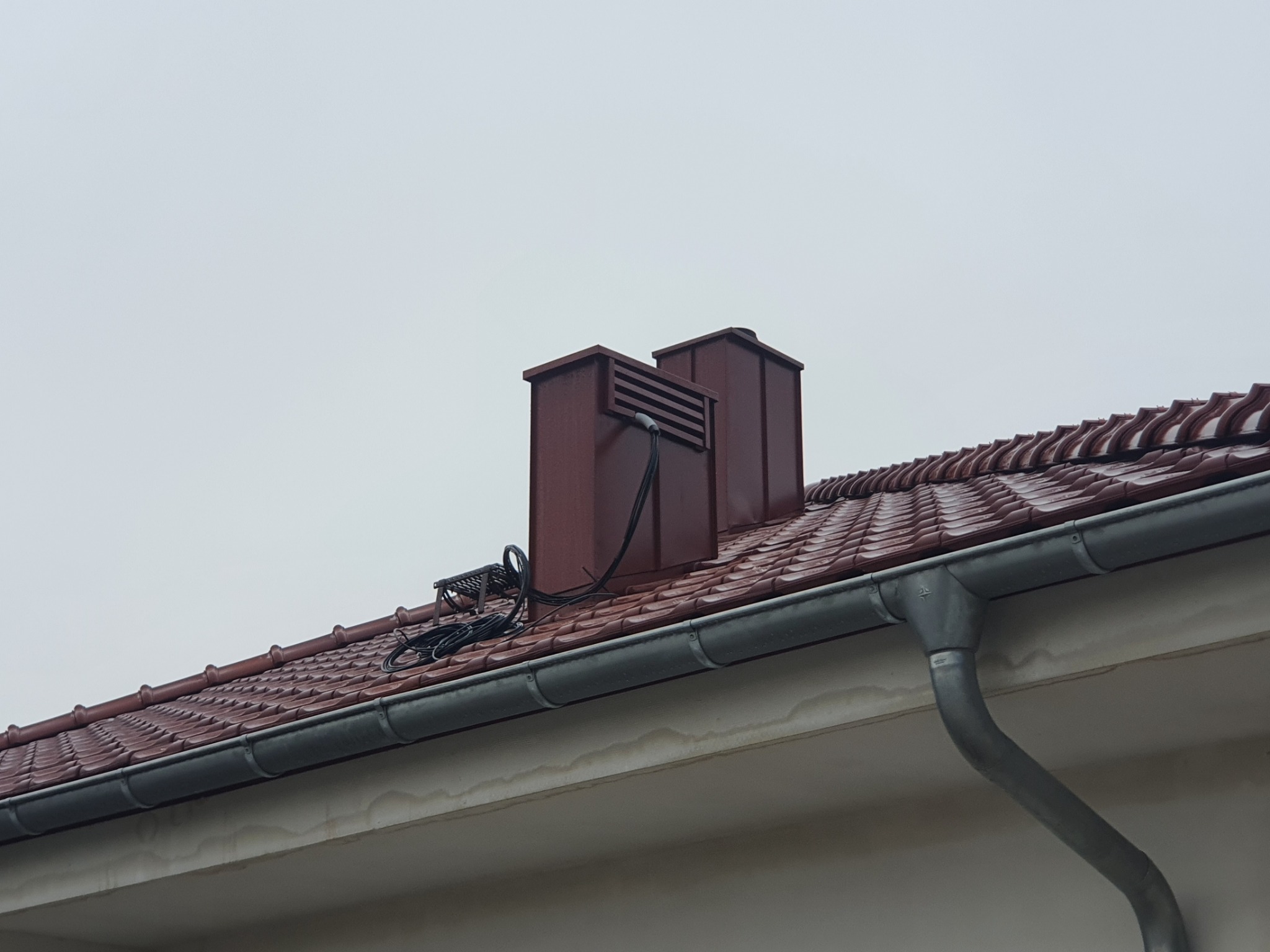 Nowy dach z brązowej dachówki ceramicznej, widoczne dwa kominy wentylacyjne w tym samym kolorze, szara rynna i kable instalacji odgromowej. Na elewacji ślady wilgoci.