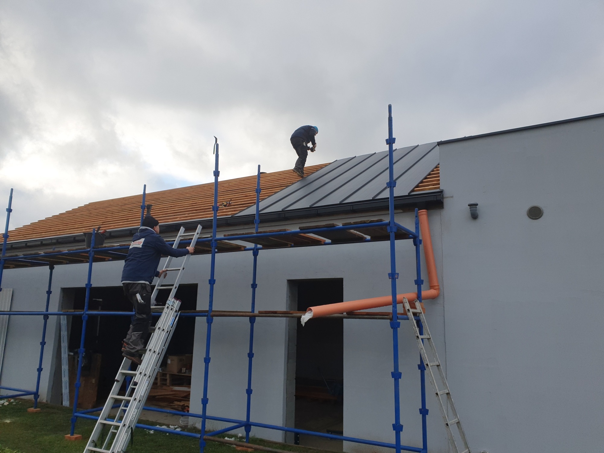 Dwóch dekarzy pracuje na rusztowaniu przy montażu blachy na dachu domu jednorodzinnego, widoczna więźba dachowa i częściowo ułożona blacha w kolorze grafitowym.