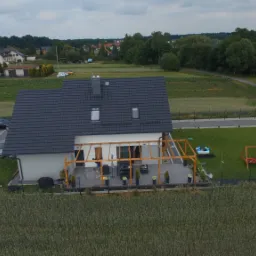 Widok z lotu ptaka na dom jednorodzinny w Zabrzu z częściowo zadaszonym tarasem z drewnianą konstrukcją, otoczony zielonym trawnikiem i polami uprawnymi.