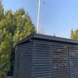 Ciemna, drewniana konstrukcja z poziomych desek z prześwitami, prawdopodobnie altana lub szopa, na tle zieleni drzew i błękitnego nieba z widocznym słupem telekomunikacyjnym.