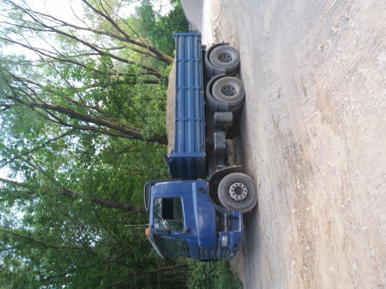 Niebieska wywrotka z ładunkiem piasku, zaparkowana na polnej drodze, otoczona zielenią drzew.