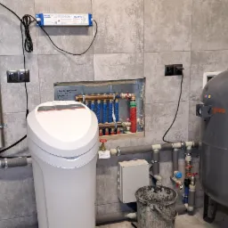 Instalacja uzdatniania wody z filtrem, lampą UV i zbiornikiem hydroforowym w kotłowni z szarymi płytkami, widoczny również rozdzielacz ogrzewania podłogowego i wiadro po zaprawie.