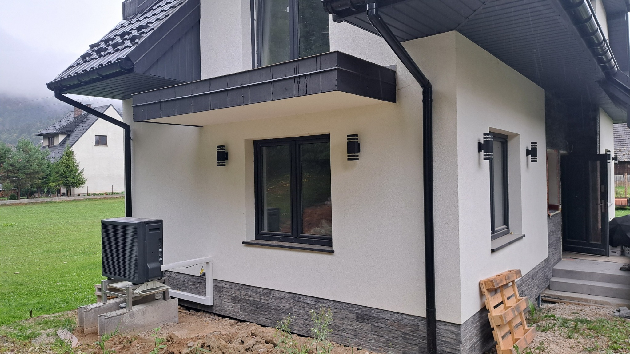 Zewnętrzna jednostka pompy ciepła zamontowana przy ścianie nowoczesnego domu z elewacją w kolorze złamanej bieli i czarnymi detalami, widoczne okno, lampy zewnętrzne, rynna i fragment palety...