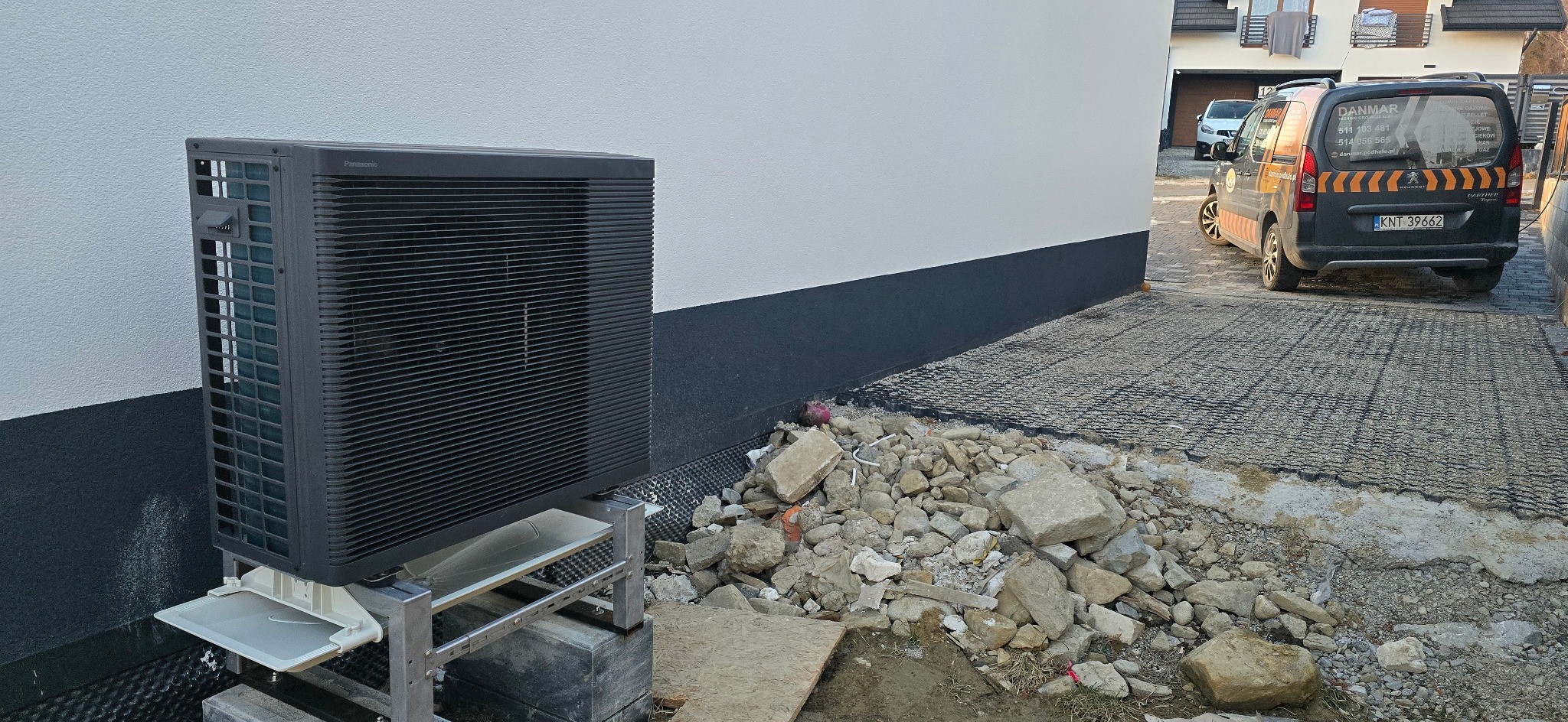 Czarna pompa ciepła Panasonic na aluminiowym stelażu, widoczna na tle białej ściany budynku z ciemnym cokołem i gruzem budowlanym na podłożu, z zaparkowanym samochodem firmy w tle.