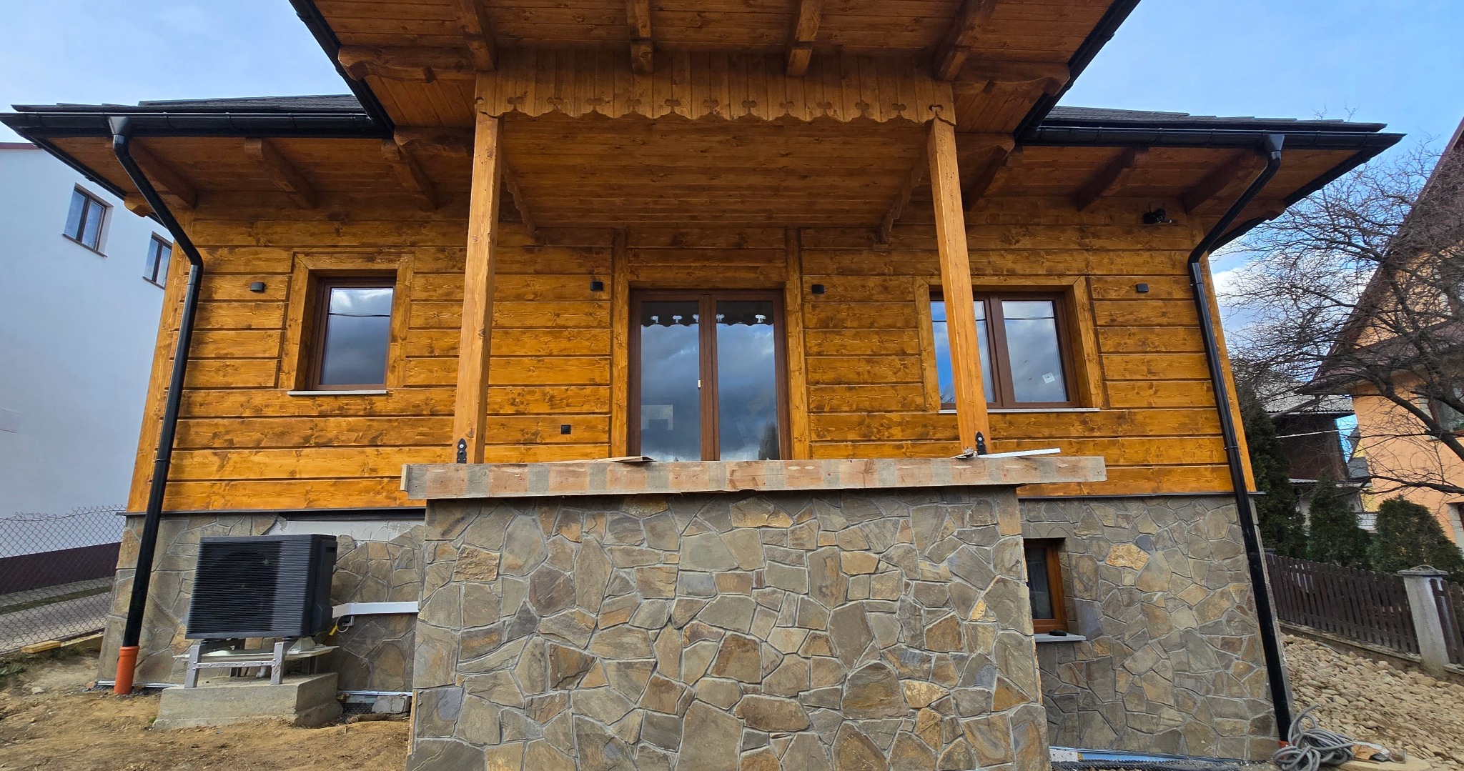 Fasada drewnianego domu z kamienną podmurówką i zamontowaną pompą ciepła na zewnątrz.