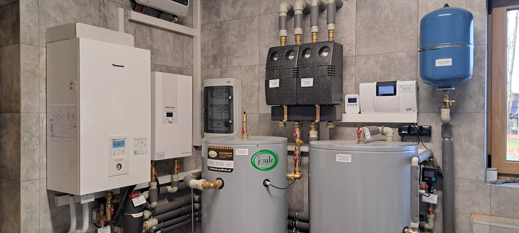 Instalacja grzewcza w kotłowni z białą pompą ciepła Panasonic, zbiornikami akumulacyjnymi Joule, rozdzielaczami w izolacji i niebieskim naczyniem przeponowym, na tle ściany z szarych płytek.