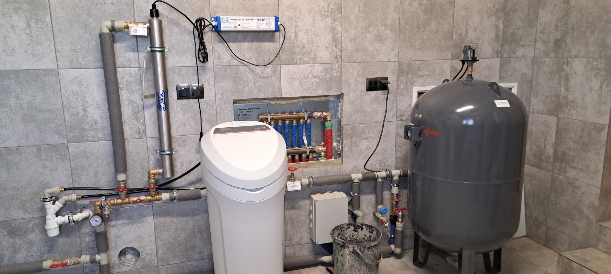 Instalacja uzdatniania wody z filtrem, lampą UV i zbiornikiem hydroforowym w kotłowni z szarymi płytkami, widoczny również rozdzielacz ogrzewania podłogowego i wiadro po zaprawie.