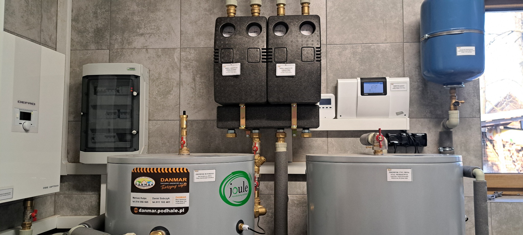Instalacja centralnego ogrzewania z dwoma zbiornikami buforowymi, rozdzielaczem z manometrami, naczyniem wzbiorczym i sterownikiem, zamontowana na ścianie z szarych płytek.