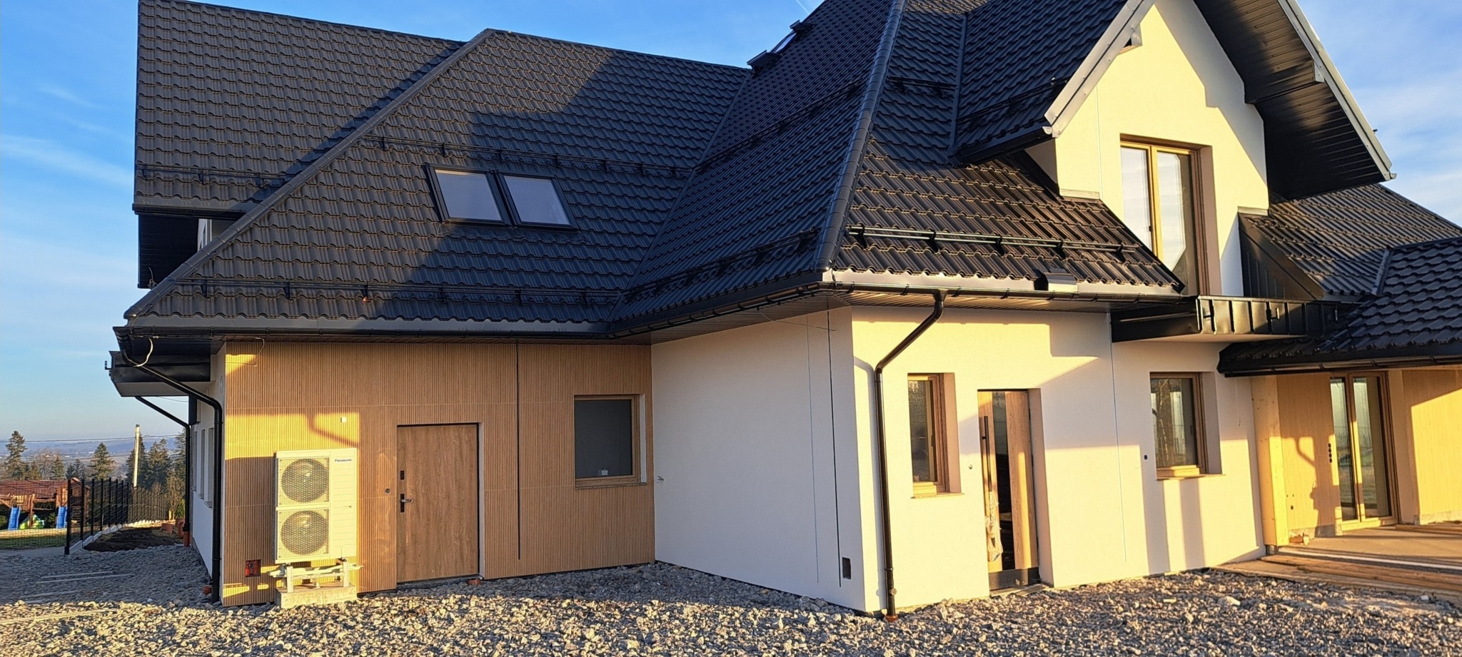 Nowoczesny dom jednorodzinny z zamontowaną pompą ciepła Panasonic na zewnątrz, widok z boku na elewację z drewnianymi panelami i grafitową dachówką.