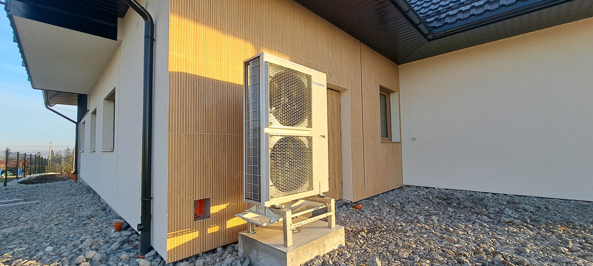 Zewnętrzna jednostka pompy ciepła Panasonic zamontowana na betonowym fundamencie przy elewacji domu z okładziną w odcieniach beżu i brązu, widok z dołu na tle kamiennego podłoża.
