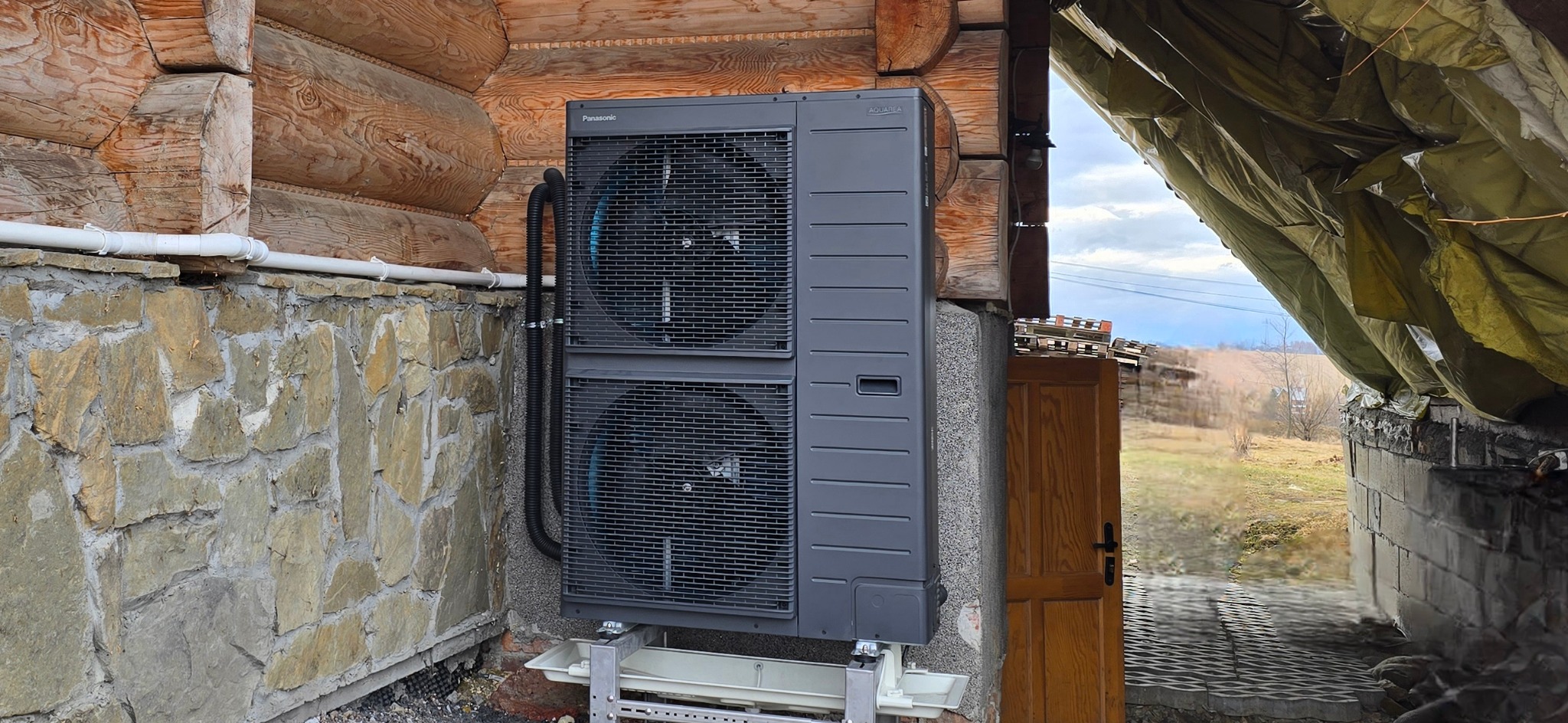 Szara, dwuwentylatorowa pompa ciepła Panasonic Aquarea zamontowana na zewnątrz drewnianego domu na kamiennej podmurówce, widoczne rury przyłączeniowe i metalowa konstrukcja wsporcza.