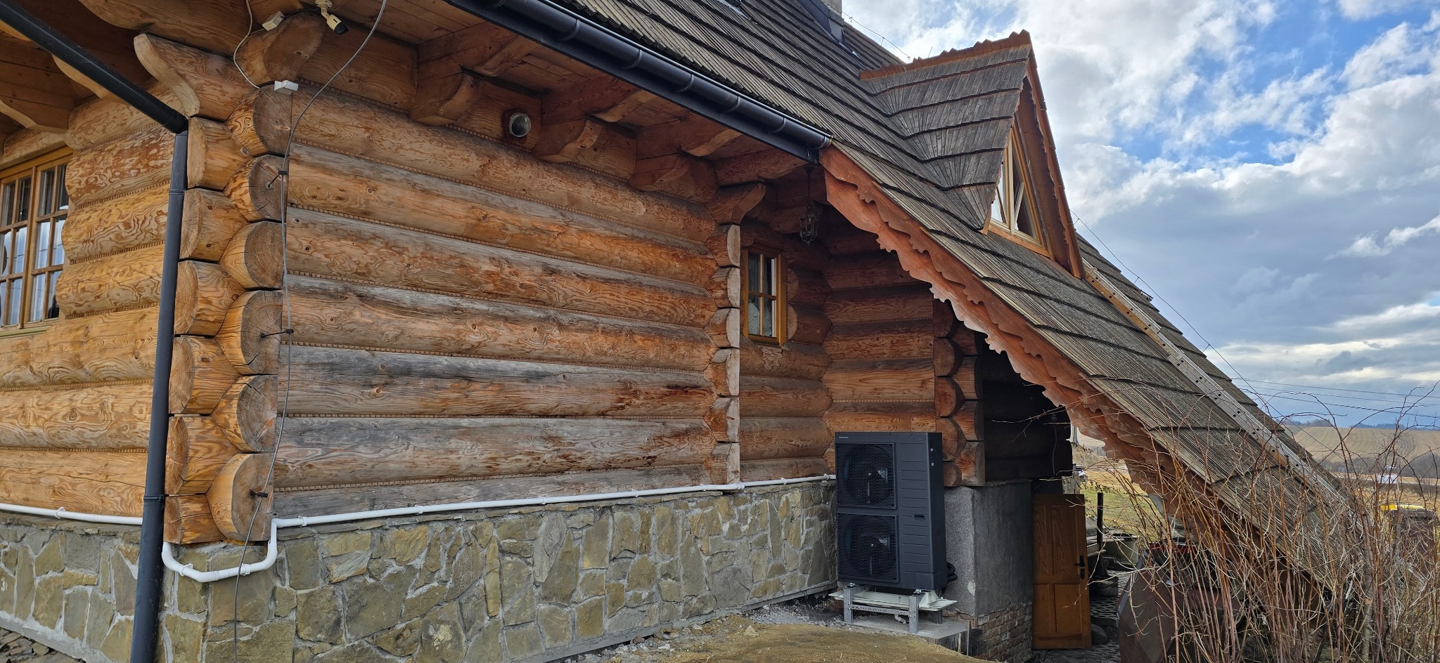 Zewnętrzna jednostka pompy ciepła zamontowana przy drewnianym domu z kamienną podmurówką, widoczna instalacja odprowadzająca skropliny.