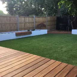 Nowoczesny ogród z drewnianym tarasem, sztuczną trawą, podwyższonymi rabatami i minimalistyczną ławką wbudowaną w mur.