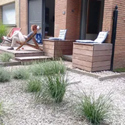 Nowoczesny ogród z białym żwirem, drewnianym tarasem i minimalistycznymi donicami. Kobieta relaksuje się na leżaku z motywem kotwicy.