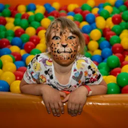 Dziewczynka z pomalowaną twarzą w kształcie pyszczka lwa, oparta o brzeg basenu wypełnionego kolorowymi piłeczkami.