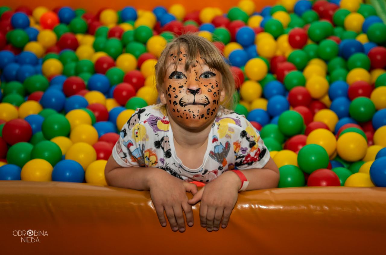 Dziewczynka z pomalowaną twarzą w kształcie pyszczka lwa, oparta o brzeg basenu wypełnionego kolorowymi piłeczkami.