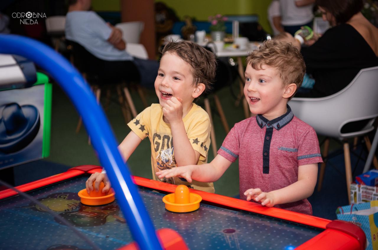Dwóch uśmiechniętych chłopców gra w air hockey, jeden w żółtej koszulce z motywem tygrysa, drugi w bordowej koszulce z granatowym kołnierzykiem, w tle siedzący ludzie i prezenty.