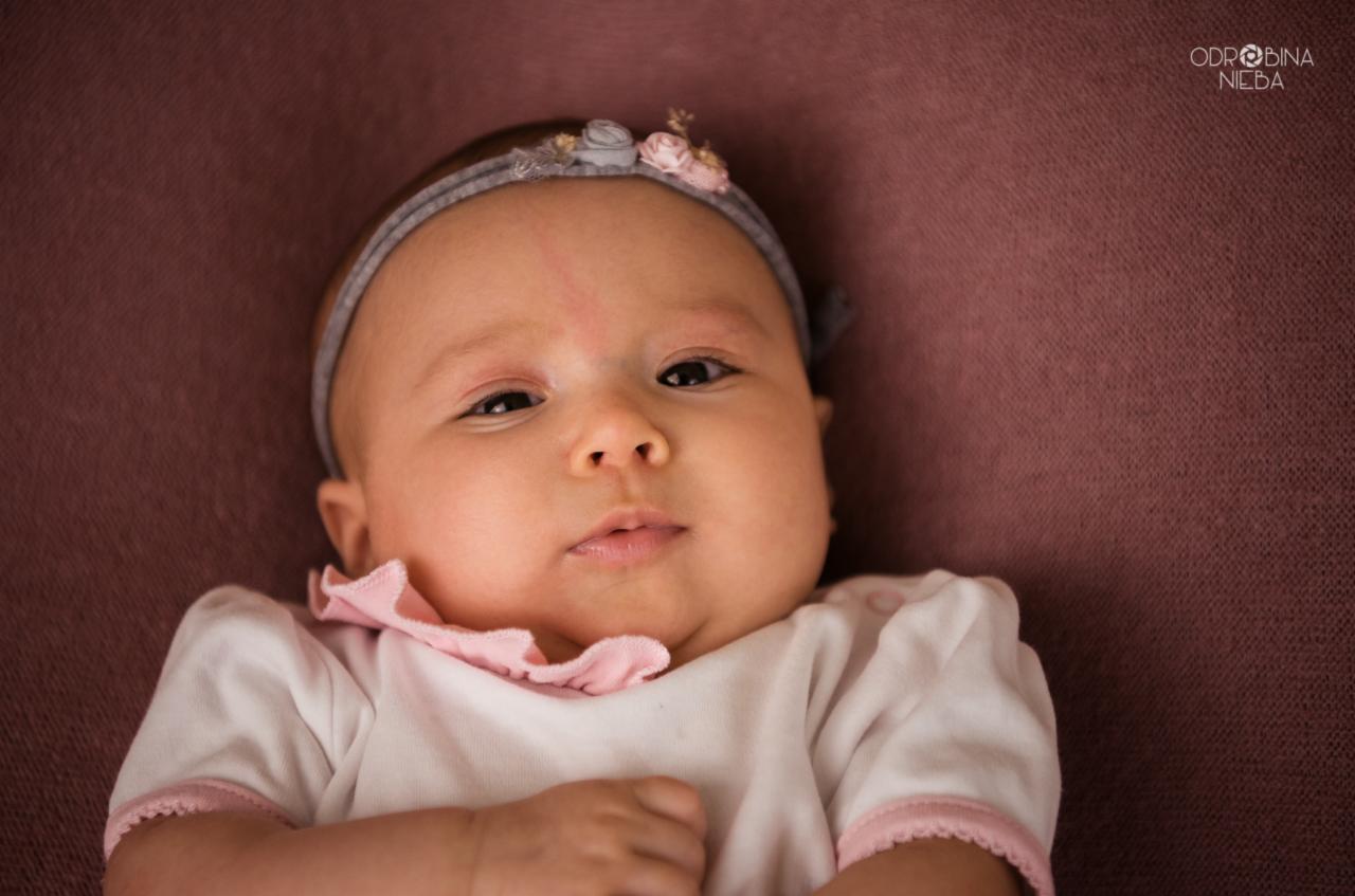 Portret niemowlęcia w białym ubranku z różowym wykończeniem i opaską z kwiatkami na głowie, leżącego na brązowym tle.