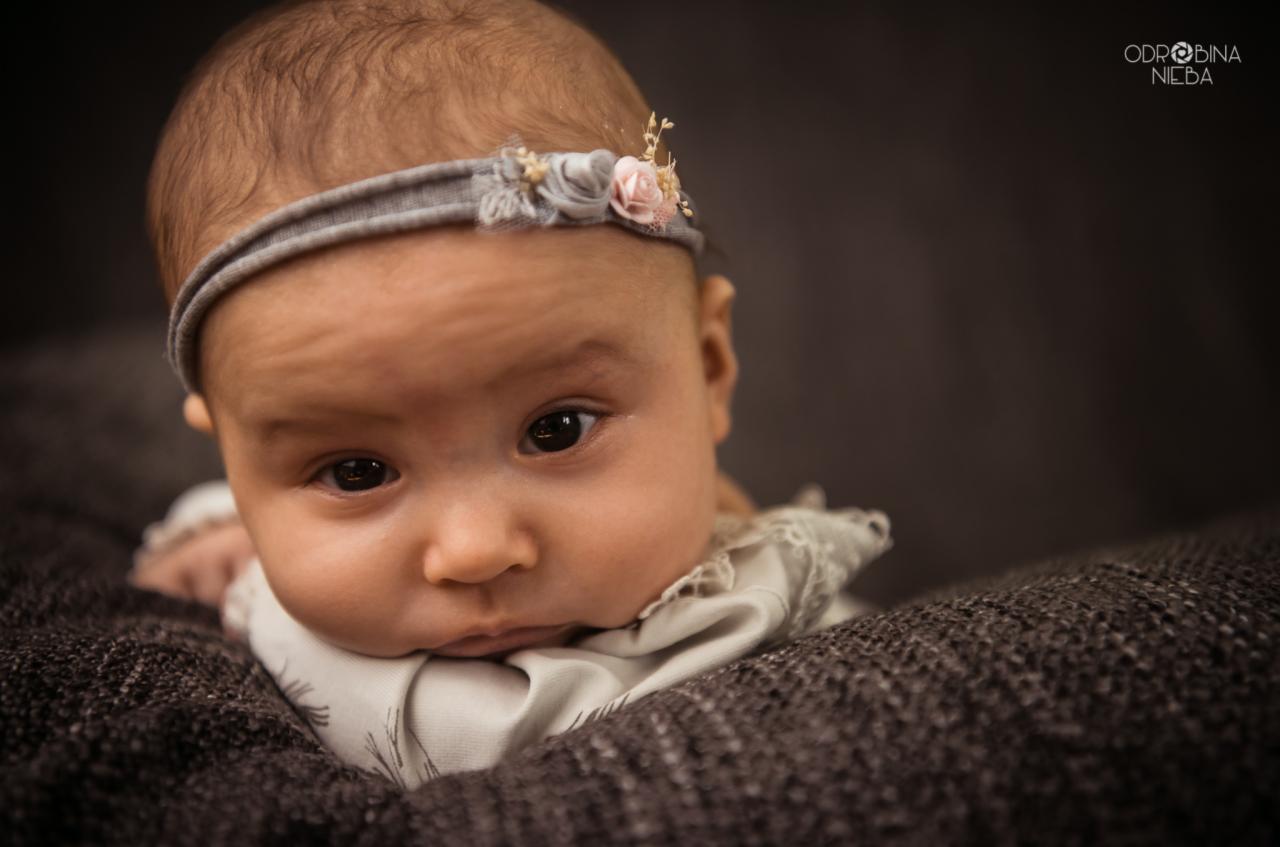Portret noworodka leżącego na ciemnoszarym kocyku, z ozdobną opaską z kwiatami na głowie, skierowanego wzrokiem w obiektyw.