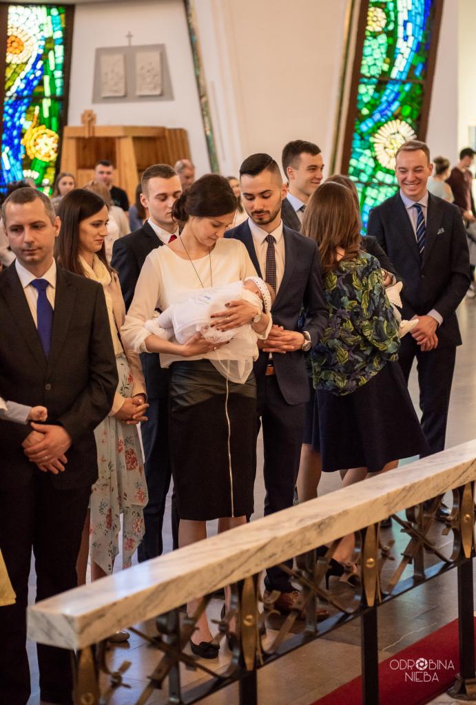 Matka trzyma nowo ochrzczone dziecko w kościele, otoczona przez rodzinę i gości, z widocznym witrażem w tle.