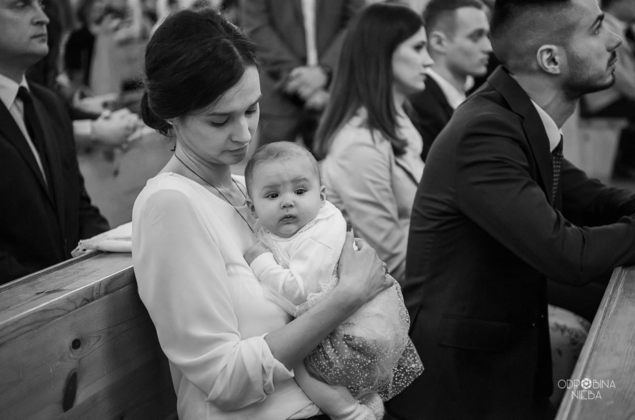 Czarno-białe zdjęcie matki trzymającej niemowlę w kościele podczas uroczystości, widoczni goście w tle.