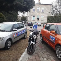 Dwa samochody i motocykl z oznaczeniami szkoły nauki jazdy zaparkowane przed budynkiem w Sierpcu. Na szybach aut naklejki z logiem i danymi kontaktowymi szkoły.