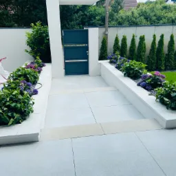 Nowoczesna aranżacja przed domem: jasnoszara kostka, podwyższone rabaty z hortensjami i czarnym grysem, minimalistyczna brama wejściowa i równo przystrzyżony trawnik w tle.