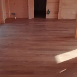 Wykończona podłoga z desek w jasnym, drewnianym domu, widoczne naturalne usłojenie i detale drewna, z fragmentem drabiny i wiszącą kurtką w tle.