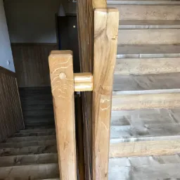 Drewniane schody z balustradą wykonane z jasnego drewna, widoczne sęki i naturalne usłojenie, fragment pomieszczenia w tle.