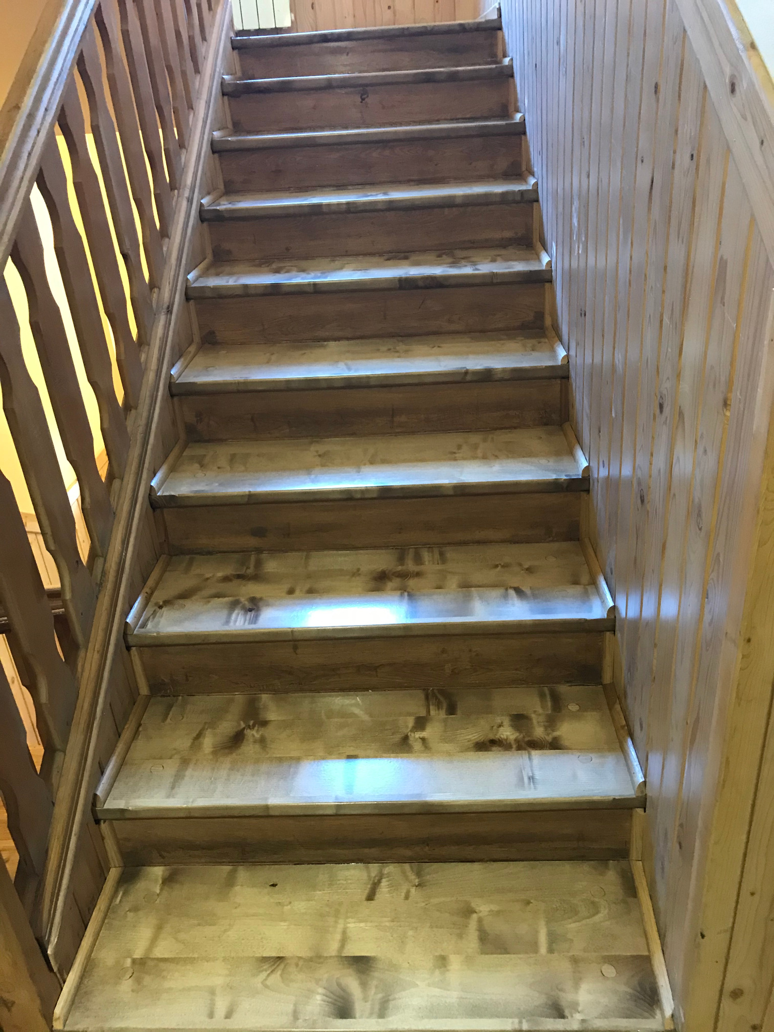 Drewniane schody z balustradą i boazerią, widok z dołu, jasne drewno z ciemniejszymi plamkami, pionowe panele ścienne.