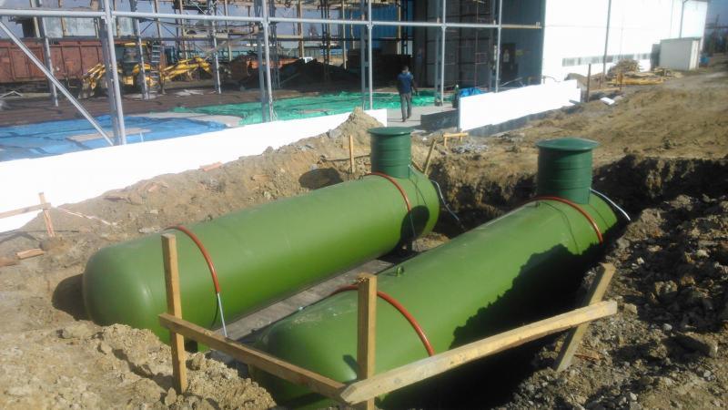Dwa zielone zbiorniki zakopane w ziemi, prawdopodobnie zbiorniki na gaz płynny, z widocznymi włazami i rurami, na tle placu budowy z konstrukcją stalową i budynkiem.