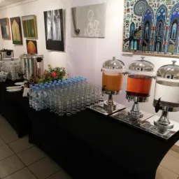 St&oacute;ł cateringowy z napojami w dyspenserach (pomarańczowy, czerwony, woda), butelkami wody, szklankami, naczyniami i kwiatami na czarnym obrusie, tło stanowią obrazy na ścianie.