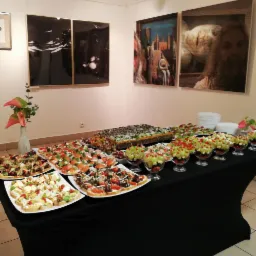 St&oacute;ł cateringowy z r&oacute;żnorodnymi przekąskami: kanapeczki z warzywami, koreczki owocowe w szklanych naczyniach, duży pasztet. W tle obrazy na ścianie i wazon z kwiatami.