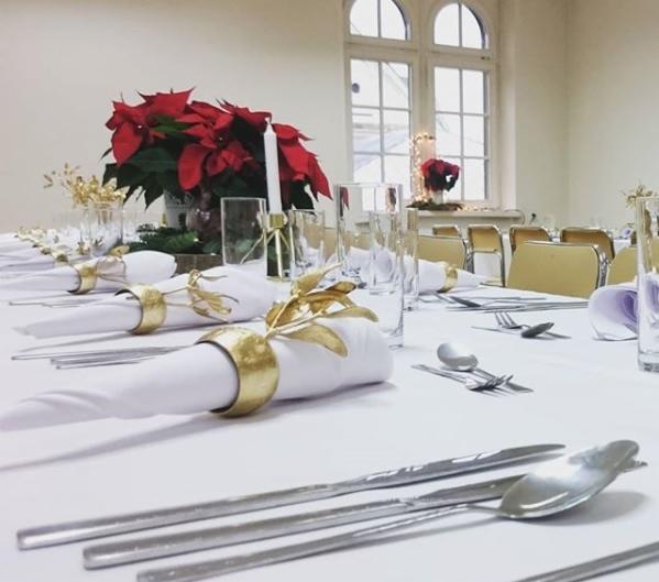 Uroczysty stół świąteczny z białym obrusem, złotymi pierścieniami na serwetkach, poinsecją i świecami w tle jasnego wnętrza z oknami.