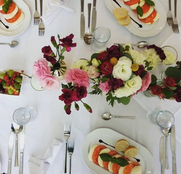 Elegancko nakryty stół z białym obrusem, dekoracjami kwiatowymi z różowych i czerwonych kwiatów, caprese z bazylią i owocami w metalowej miseczce.
