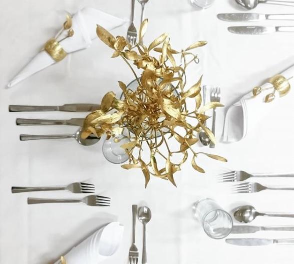 Elegancko nakryty stół z białym obrusem, złotymi dekoracjami roślinnymi w wazonie, sztućcami i serwetkami ze złotymi pierścieniami, widok z góry.