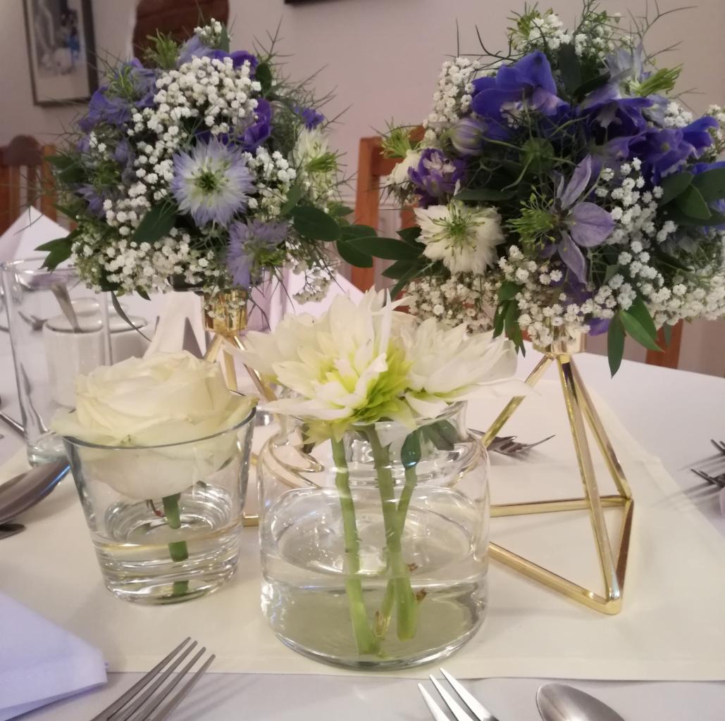 Dekoracja stołu: kompozycje z białych i fioletowych kwiatów w szklanych wazonach i geometrycznych, złotych stojakach, subtelne oświetlenie.
