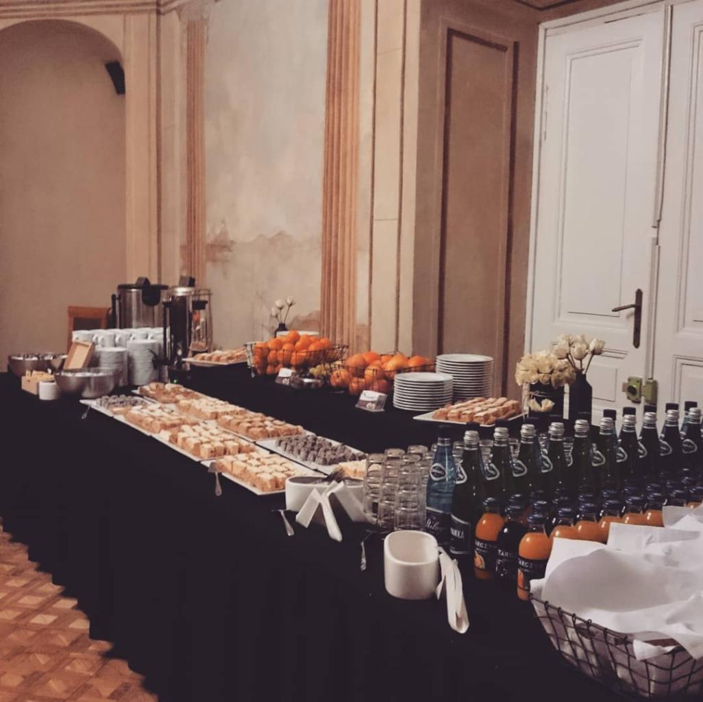 Stół cateringowy z czarnym obrusem: ciasta, owoce, napoje w butelkach i dzbanki na tle klasycznego wnętrza z wysokimi drzwiami i sztukaterią.