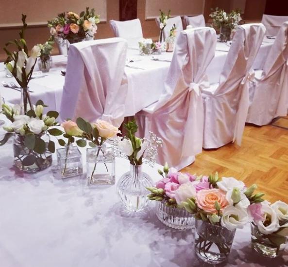 Dekoracja stołu na uroczystość: niskie wazoniki z różowymi i białymi kwiatami, białe obrusy i krzesła z białymi pokrowcami zawiązanymi na kokardę.