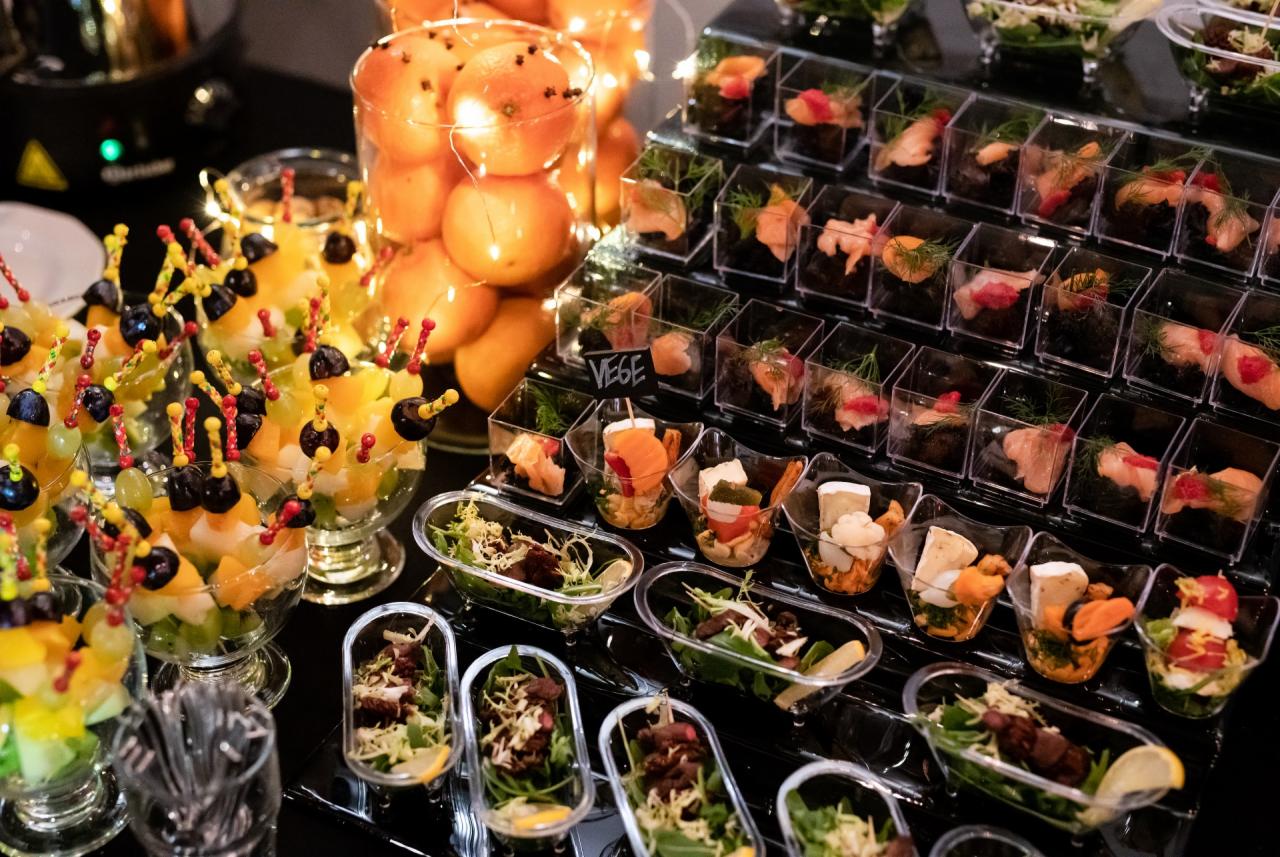 Elegancko zaaranżowany bufet z różnorodnymi przekąskami w małych naczyniach: owocowe szaszłyki, sałatki i przystawki rybne na czarnym, błyszczącym stole, pomarańcze w tle.