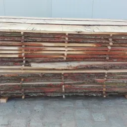 Stos tarcicy obrzynanej z widoczną korą, ułożonej warstwami na przekładkach, na zewnątrz budynku.