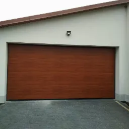 Zamknięta, segmentowa brama garażowa w kolorze drewna, zamontowana w jasnym garażu z szarym brukowanym podjazdem.