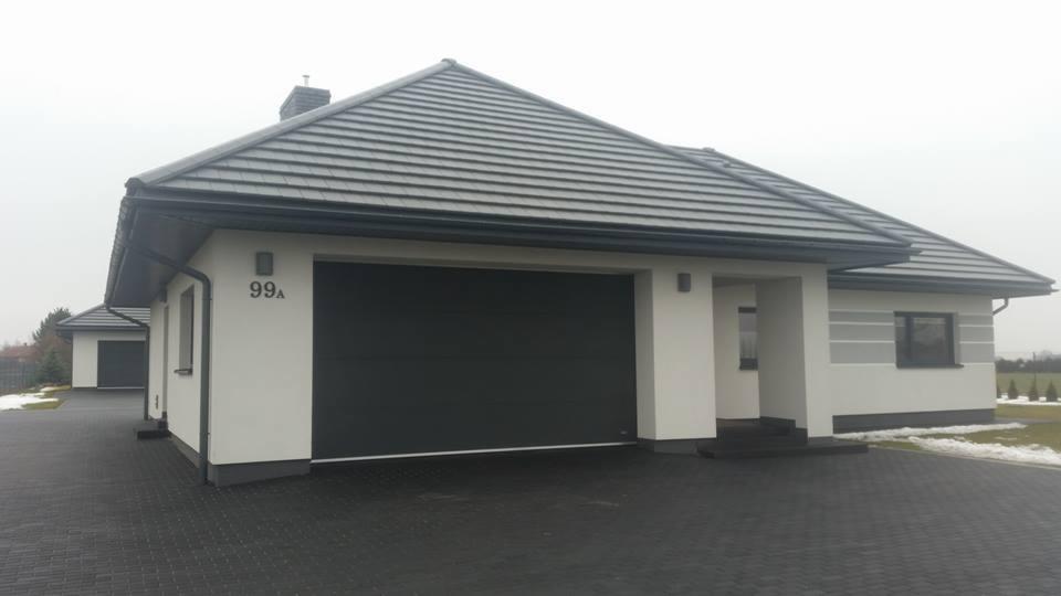 Nowoczesny dom jednorodzinny z szarą, segmentową bramą garażową, numerem domu 99A i podjazdem z kostki brukowej w pochmurny dzień.