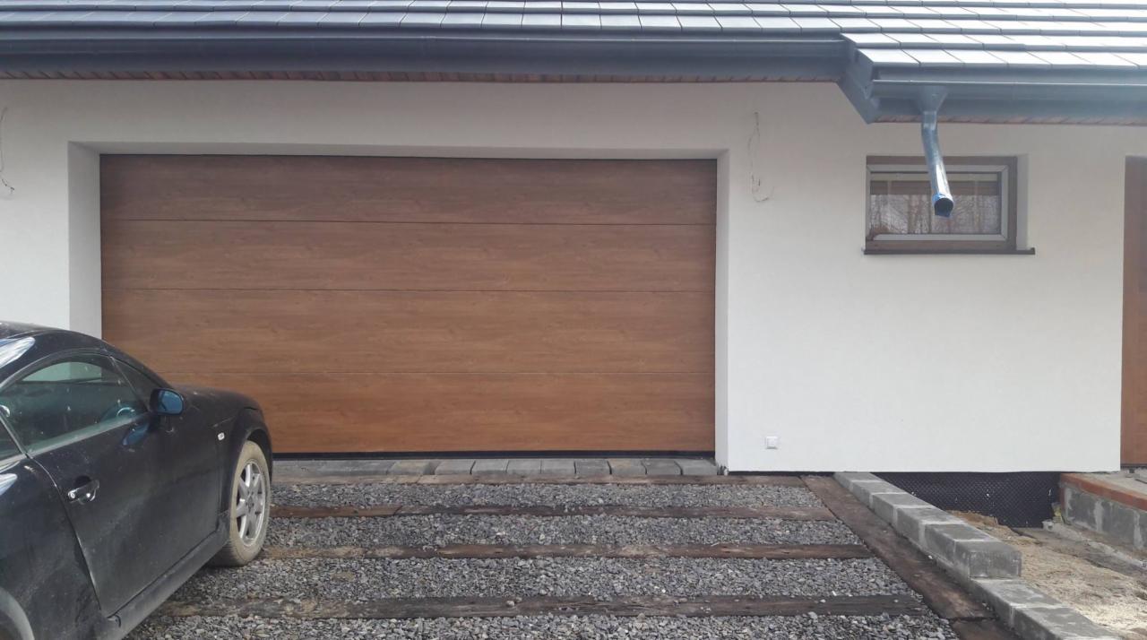 Zewnętrzny widok na segmentową bramę garażową w kolorze drewna, z widocznym fragmentem czarnego samochodu i szarej nawierzchni przed garażem.