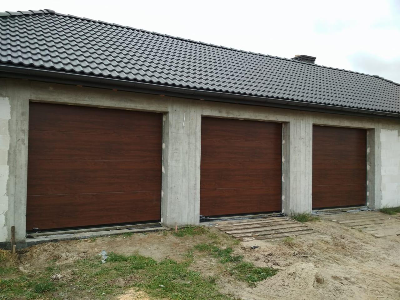 Trzy brązowe segmentowe drzwi w surowym garażu z betonowymi ścianami i czarnym dachem z dachówki. W tle pochmurne niebo.