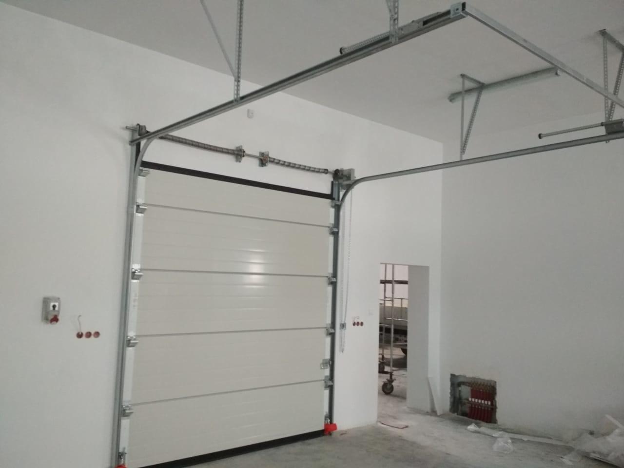 Zamknięta segmentowa brama garażowa z widocznym systemem prowadnic pod sufitem, zainstalowana w Lubartowie w nowym, surowym garażu z widocznymi instalacjami elektrycznymi i grzewczymi.