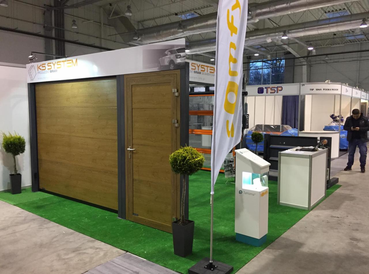 Ekspozycja brązowej segmentowej bramy garażowej z drzwiami bocznymi na stoisku targowym, z logo producenta KS System i flagą reklamową Somfy, na sztucznej trawie.