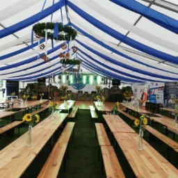 Wnętrze namiotu imprezowego z drewnianymi stołami i ławkami, dekoracjami z girland i preclami podwieszonymi pod sufitem w biało-niebieskie pasy, słoneczniki w wazonach na stołach, zielona trawa...