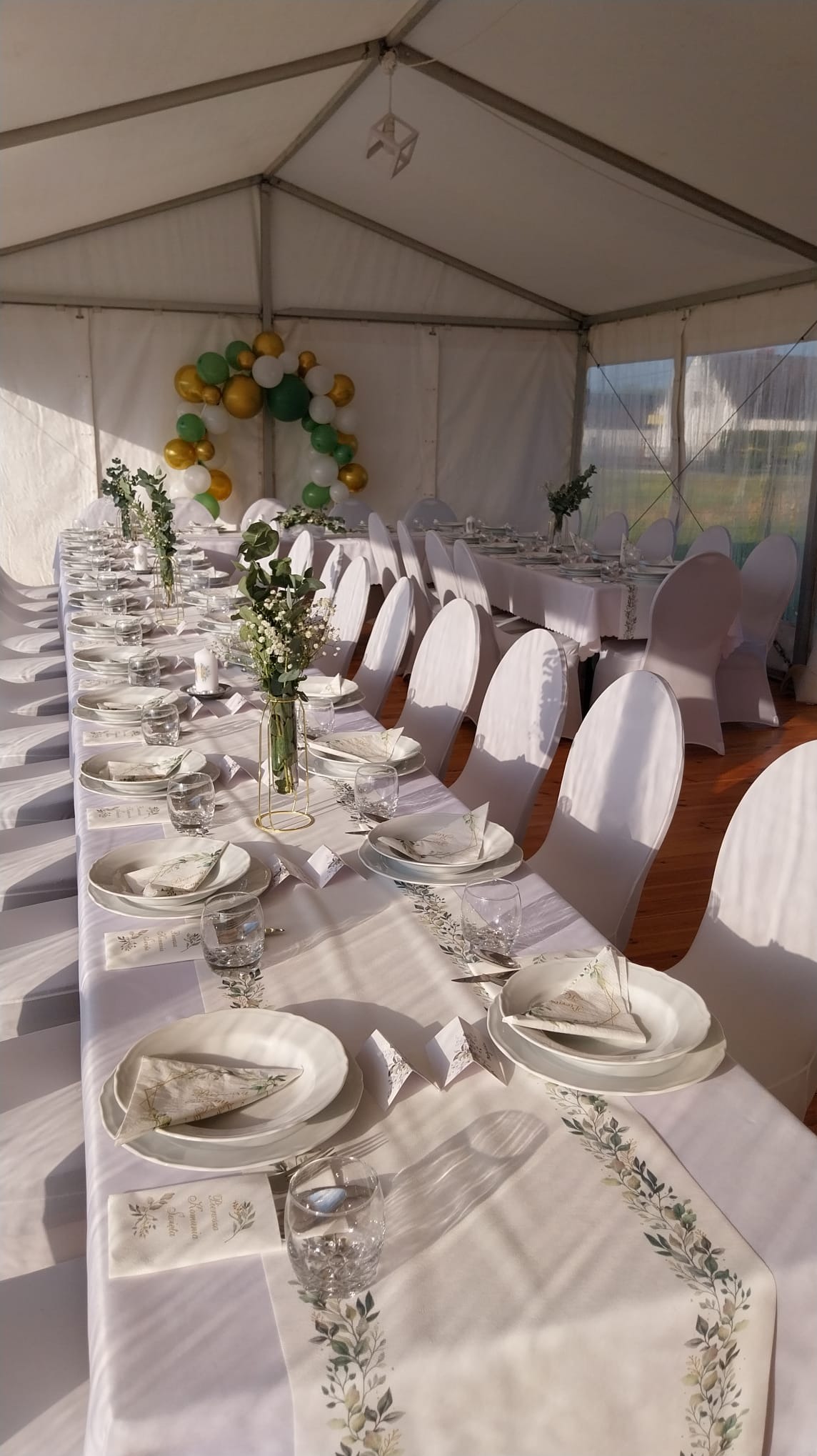Uroczysty stół weselny nakryty białym obrusem z motywem roślinnym w namiocie, dekoracje z zielonych i złotych balonów w tle.